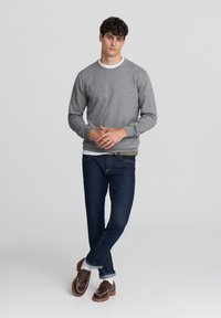 Felpa grigia con colletto e polsini a coste, indossata sopra una camicia bianca. Abbinata a jeans blu scuro e scarpe marroni con suole bianche.