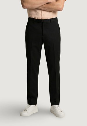 Mężczyzna w czarnych, szytych na miarę spodniach, beżowej koszuli z krótkim rękawem włożonej do środka oraz białych trampkach, stojący z założonymi ramionami.