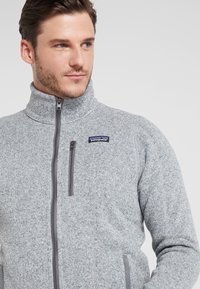 Gray fleece jacket with a high collar, full zip closure, and a left chest pocket. Features a textured pattern and gray hardware accents.