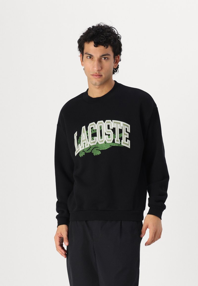 Sudadera negra con escote redondo, que presenta un gran logo "LACOSTE" en verde y blanco y un diseño de cocodrilo impreso en la parte delantera.