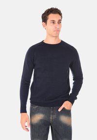 Maglione a maglia blu navy con collo a giro, polsini e orlo a coste. Indossato con jeans blu sbiaditi, evidenziando un fitting rilassato e un tessuto testurizzato.