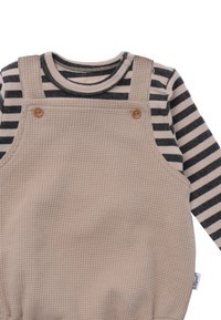 Beige strukturierte Latzhose mit Holzknöpfen, über einem schwarz-weiß gestreiften Langarmshirt getragen. Weicher, gemütlicher Stoff.