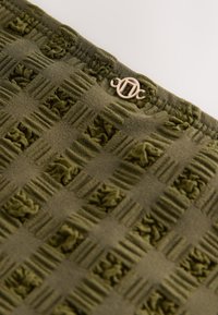 Tissu vert olive avec un motif damier texturé et un petit logo métallique rond doré fixé près du bord.