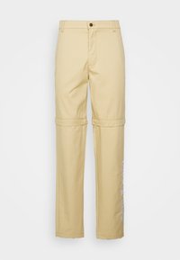 Pantalons droits beige avec une fermeture à bouton, dotés d'une couture pour se transformer en version roulée et d'un texte imprimé le long de la jambe gauche.