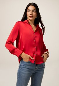 Blouse en satin rouge avec un boutonnage sur le devant, des manches longues et un col. Portée avec un jean ajusté bleu, mettant en valeur une texture lisse.