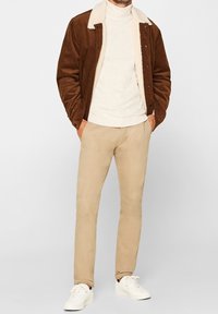 Veste en velours côtelé marron avec col en fausse peau de mouton beige, superposée sur un col roulé beige clair, associée à un pantalon slim beige clair et des baskets blanches.