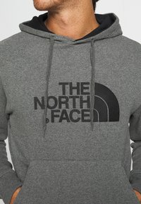 Felpa grigia con cappuccio e logo nero caratterizzato da un design a semicerchio. Realizzata in materiale morbido, presenta una tasca frontale e cordini.