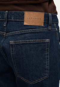 Σκούρο μπλε τζιν με πορτοκαλί ραφές και καφέ δερμάτινο ταμπελάκι με την ένδειξη "TOPSHOP" στη πίσω ζώνη της μέσης.