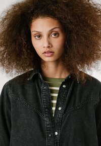 Juodas džinsinis švarkas su spaudėmis, vakarietiško stiliaus kaklas, nustatyta tekstūra, dėvimas ant žaliai ir baltai dryžuoto marškinėlių.