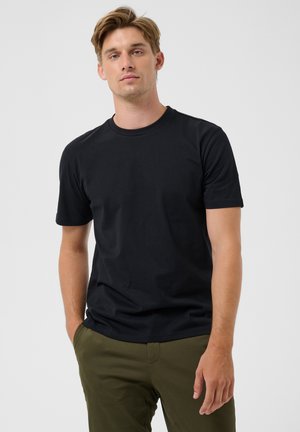 Joven con cabello castaño claro que lleva una camiseta negra de manga corta y pantalones verde oliva, de pie frente a un fondo claro y liso.