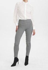 Bílá krajková blůza s vysokým límcem a zapínáním na knoflíky, která je spárována s černobílými kostkovanými kalhotami s vysokým pasem a černými kotníkovými botami.