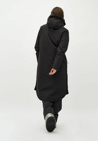 Zwarte lange jas met een capuchon, gemaakt van een glad materiaal. Heeft zijsplitten, lange mouwen en wordt gedragen over een gevoerde zwarte broek en laarzen.