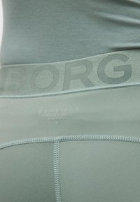 Κοντινό πλάνο στη ζώνη μέσης ενός ανοιχτοπράσινου αθλητικού σορτς με μερικώς ορατό το όνομα της μάρκας και λεπτομερείς ραφές στο ύφασμα.