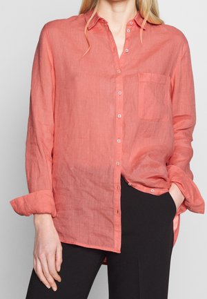 Femme portant une chemise ample corail à manches longues boutonnée, avec les poignets retroussés, une main dans la poche d’un pantalon noir, sur un fond clair uni.