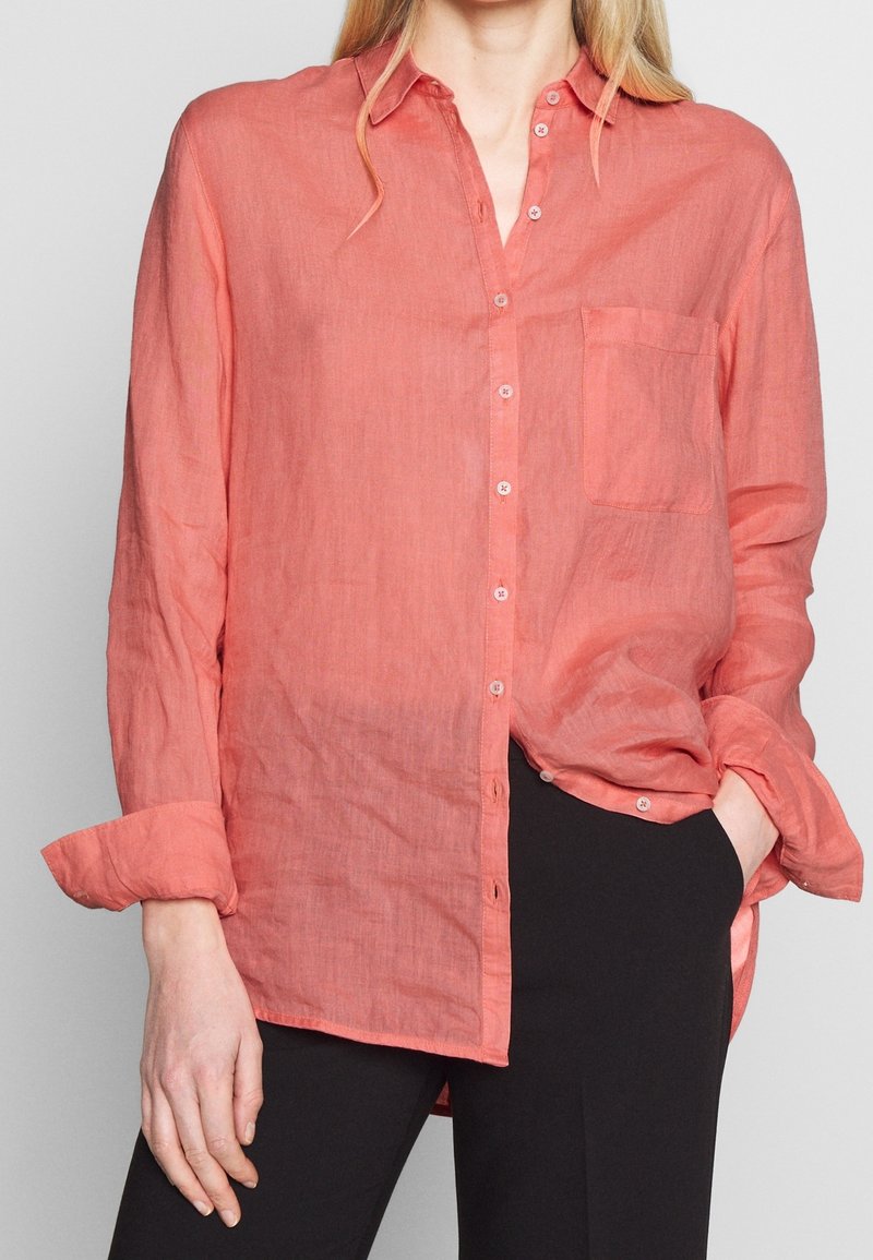 Femme portant une chemise ample corail à manches longues boutonnée, avec les poignets retroussés, une main dans la poche d’un pantalon noir, sur un fond clair uni.