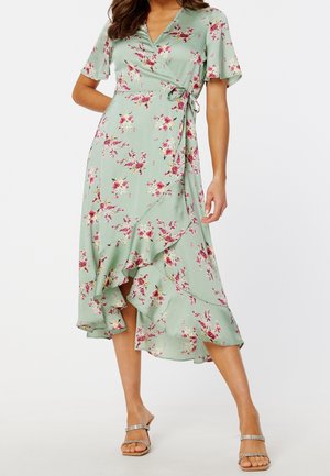Femme portant une robe portefeuille à fleurs vert clair avec un ourlet asymétrique à volants, des sandales à talons hauts à brides, debout, une main sur la hanche.