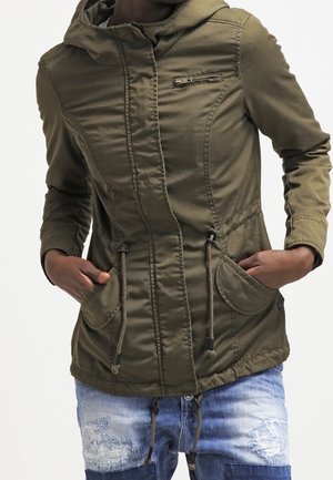 Person trägt eine olivgrüne Kapuzenjacke mit Frontreißverschluss und Taschen, Hände in den Taschen, kombiniert mit abgetragenen blauen Jeans.