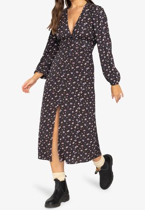 Frau trägt ein schwarzes, geblümtes Midi-Kleid mit langen Ärmeln und Frontschlitz, kombiniert mit cremefarbenen Socken und schwarzen Stiefeletten, posiert beim Gehen.