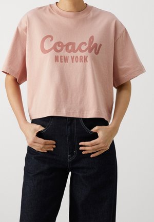 Person trägt ein pinkes, kurzes T-Shirt mit dem Schriftzug „Coach New York“ und dunkle, hoch taillierte Jeans, Hände in den vorderen Taschen ruhend.