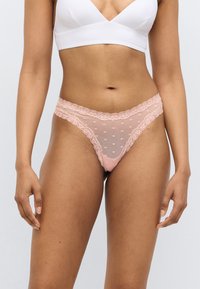 String en dentelle rose clair avec panneaux en mesh, présentant de petits motifs en forme de cœur et une bordure en dentelle décorative sur les bords. Texture douce et design délicat.
