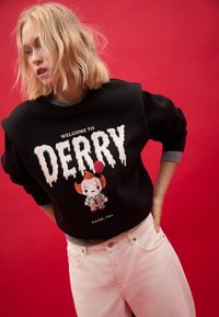 Felpa nera con la scritta "WELCOME TO DERRY" in lettere bianche che sembrano gocciolare, con un personaggio dei cartoni colorato che tiene un palloncino. Jeans rosa chiaro.