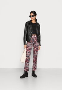 Jaqueta de motoqueiro em couro preto sobre um top preto curto, combinada com calças de padrão abstrato multicolorido, botas de cano curto pretas e uma bolsa de ombro creme.