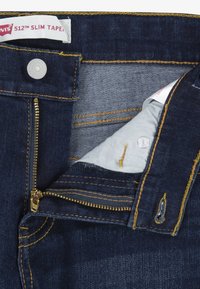 Jeans de mezclilla azul oscuro con cremallera, cosidos con hilo amarillo, y una etiqueta blanca que dice "Levi's 512 Slim Taper."