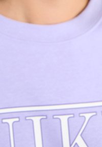 Liliowy T-shirt o gładkiej fakturze i okrągłym dekolcie, z widocznym białym wzorem na przodzie.