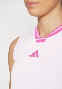 Bijela atletska majica bez rukava s ružičastim detaljima. Ima okrugli izrez i teksturirani materijal. Na prsima prikazuje ružičasti adidas logo.