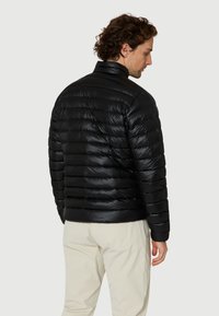 Mand med krøllet hår iført en skinnende sort polstret jakke og lysebeige bukser, vist fra ryggen mod en enkel baggrund.