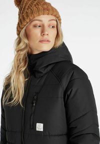 Sort polstret jakke med høj hals og hætte. Har en lynlåslomme på brystet og et logo-patch. Bæres med en strikket tan beanie.