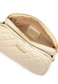 Borsa a tracolla in pelle beige trapuntata con hardware dorato. Presenta due scomparti con zip e un’etichetta logo interna. Esterno testurizzato.