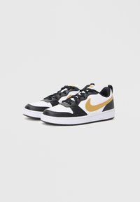 Une paire de baskets basses avec des accents blancs, noirs et dorés, des lacets noirs et un swoosh Nike doré sur un fond clair.