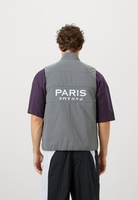 Gray puffer vest with high collar and large white "PARIS" text on the back, paired with a purple short-sleeve shirt and black pants.
