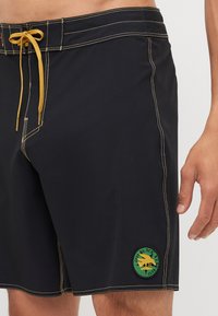 Čierne plávacie šortky so žltým prešívaním, elastický pás so šnúrkou a okrúhla zelená nášivka s logom na ľavom stehne.