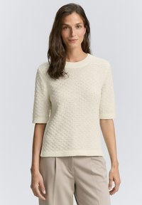 Maglione a maniche corte color crema con motivo lavorato a maglia, colletto e polsini a coste, abbinato a pantaloni beige a vita alta.