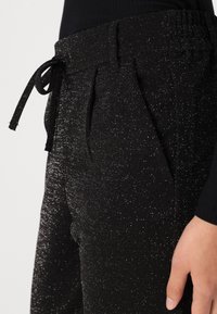 Pantalons noirs avec une taille à cordon, dotés d'un tissu texturé qui scintille avec des fils argentés. Design plissé à l'avant.