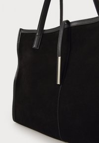 Sac fourre-tout en suède noir avec des accents en cuir lisse et des anses élégantes. Comprend un détail de logo métallique et un design structuré et minimaliste.