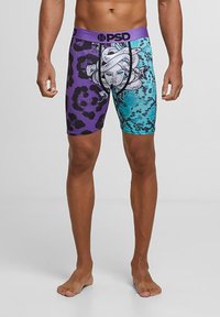 Männliches Model trägt lila-blaue Boxershorts mit schwarzem Schlangen- und Gesichtsgrafik und Tiermuster, steht barfuß auf einfarbigem Hintergrund.