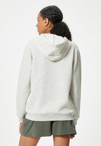Sudadera gris claro con un bolsillo tipo canguro, puños y dobladillo acanalados. Presenta una capucha ajustable y una textura cepillada. Combinada con pantalones cortos verdes.