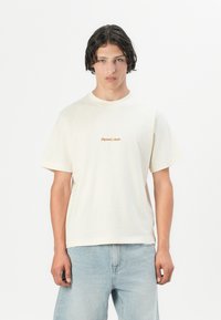 Tricou din bumbac de culoare crem, cu mâneci scurte, având un logo brodat subtil în nuanțe de portocalie. Construit cu un croi lejer și decolteu rotund.