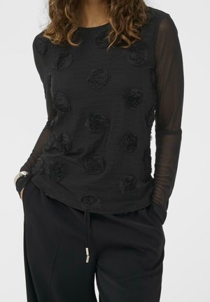 Frau trägt ein schwarzes Langarmtop mit durchsichtigen Ärmeln und erhöhten Blumenapplikationen, kombiniert mit schwarzen Hosen und einem silbernen Armband.