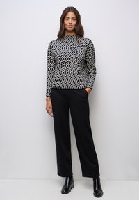 Der Pullover hat ein schwarz-weißes geometrisches Muster, einen hohen Kragen, lange Ärmel und wird mit schwarzen, weit geschnittenen Hosen sowie schwarzen Stiefeletten kombiniert.