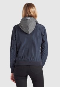Mörkblå bomberjacka med en grå huva, elastiska ärmslut och midjeresår. Har ett slätt, blankt material och en logotagg på huven.