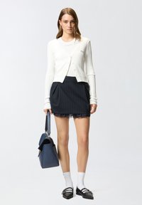 Bijela rebrasta cardigan jakna s gumbima preko tamnoplave plisirane suknje, u kombinaciji s crnim balerinkama i plavom torbom. Bijele anklet čarape upotpunjuju izgled.