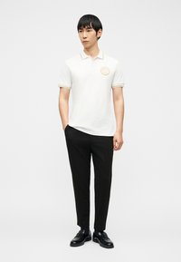 Άνδρας που φοράει λευκό πόλο με κοντά μανίκια, χρυσή διακόσμηση και έμβλημα, μαύρο παντελόνι και μαύρα παπούτσια, στέκεται με το ένα χέρι στην τσέπη.