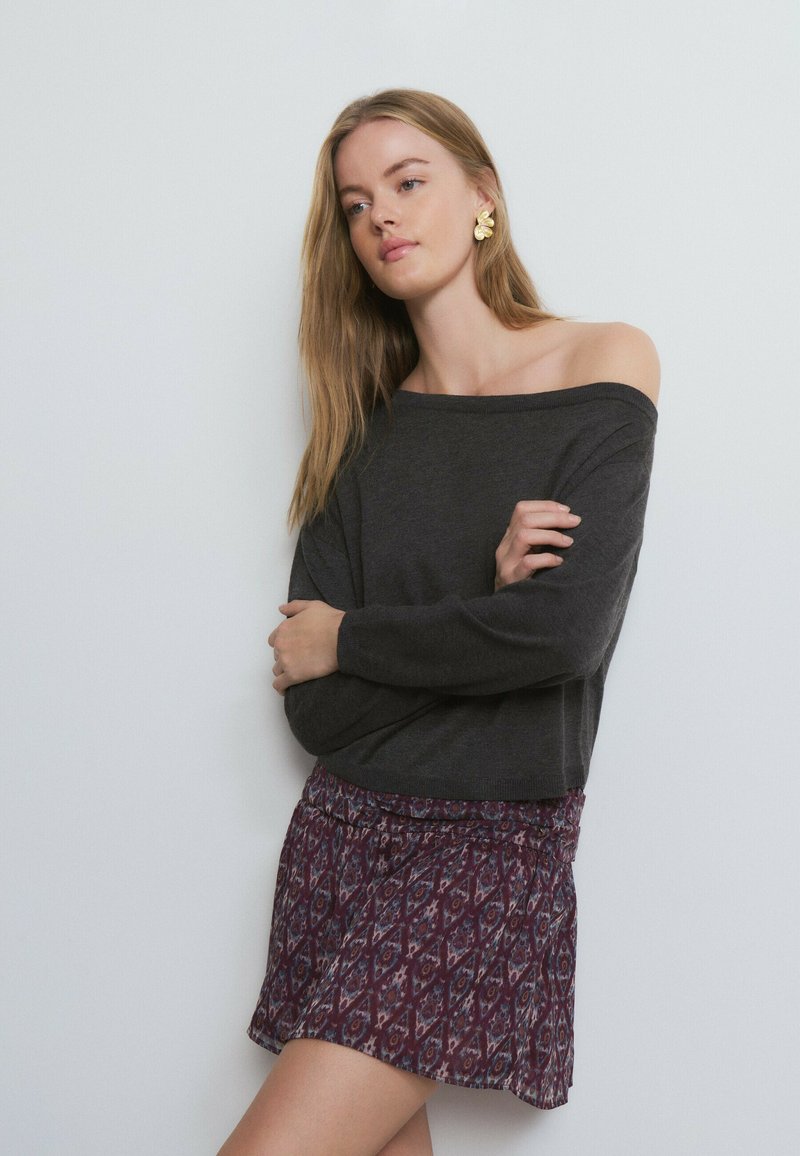 Suéter gris oscuro con hombros descubiertos combinado con una falda corta morada con estampados geométricos. Modelo posando frente a un fondo claro.