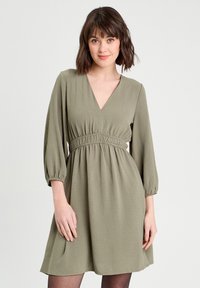 Robe vert olive avec un décolleté en V, taille élastique et manches bouffantes trois quarts. Fabriquée en tissu léger avec une texture douce.