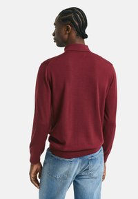 Le pull polo à manches longues bordeaux présente un col et un ourlet côtelé, fabriqué en tissu doux, avec une texture subtile et une coupe ajustée.