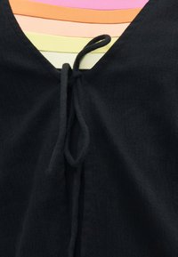 Μαύρο αμάνικο κορμάκι με V-λαιμόκοψη και λεπτομέρεια δεσίματος, φορεμένο πάνω από μπλουζάκια σε ροζ, πορτοκαλί, κίτρινο και ανοιχτό πράσινο χρώμα.